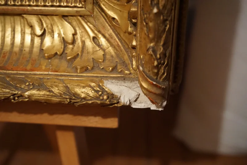 Close-up of carved gilt frame corner with visible chip and exposed gesso, condition detail
