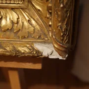 Close-up of carved gilt frame corner with visible chip and exposed gesso, condition detail